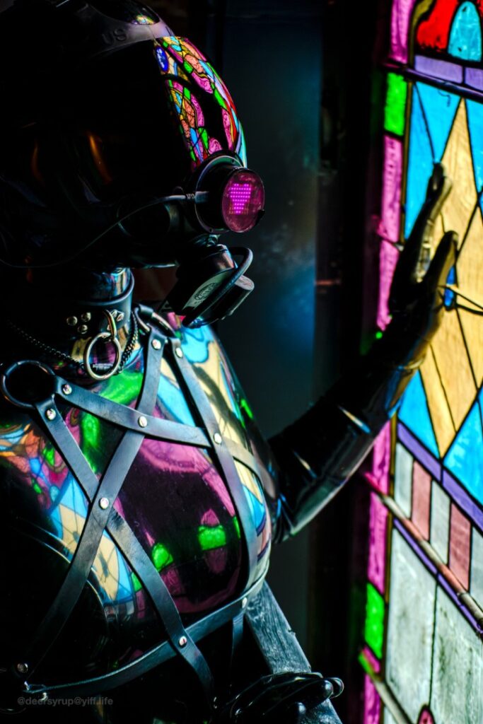 a figure clad in glossy black latex and a black full-face respirator mask. it is holding its hand up to a vibrant stain glass window, the light filtering through reflecting upon and illuminating their otherwise inkyblack silhouette. the saturated, chromatic reflections are framed by a black leather harness, forming a pentacle of matte black against their radiantly reflective form.