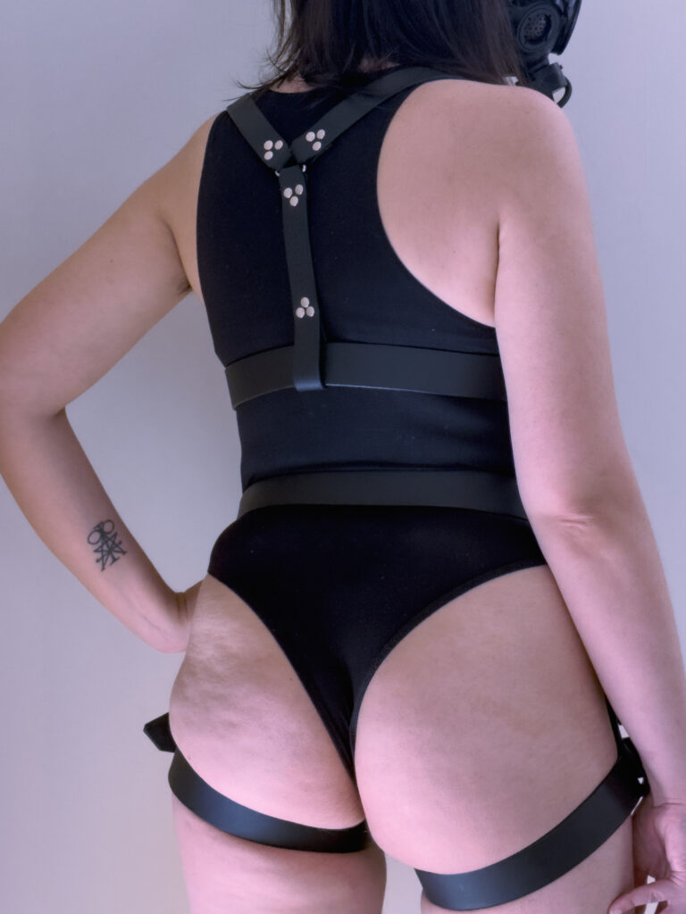 photograph from behind of a figure wearing a full-face tinted-visor respirator, modelling a wide, black leather chest- and waist-harness set. they are standing with relaxed posture, with one hand resting on their hip and the other at their side.